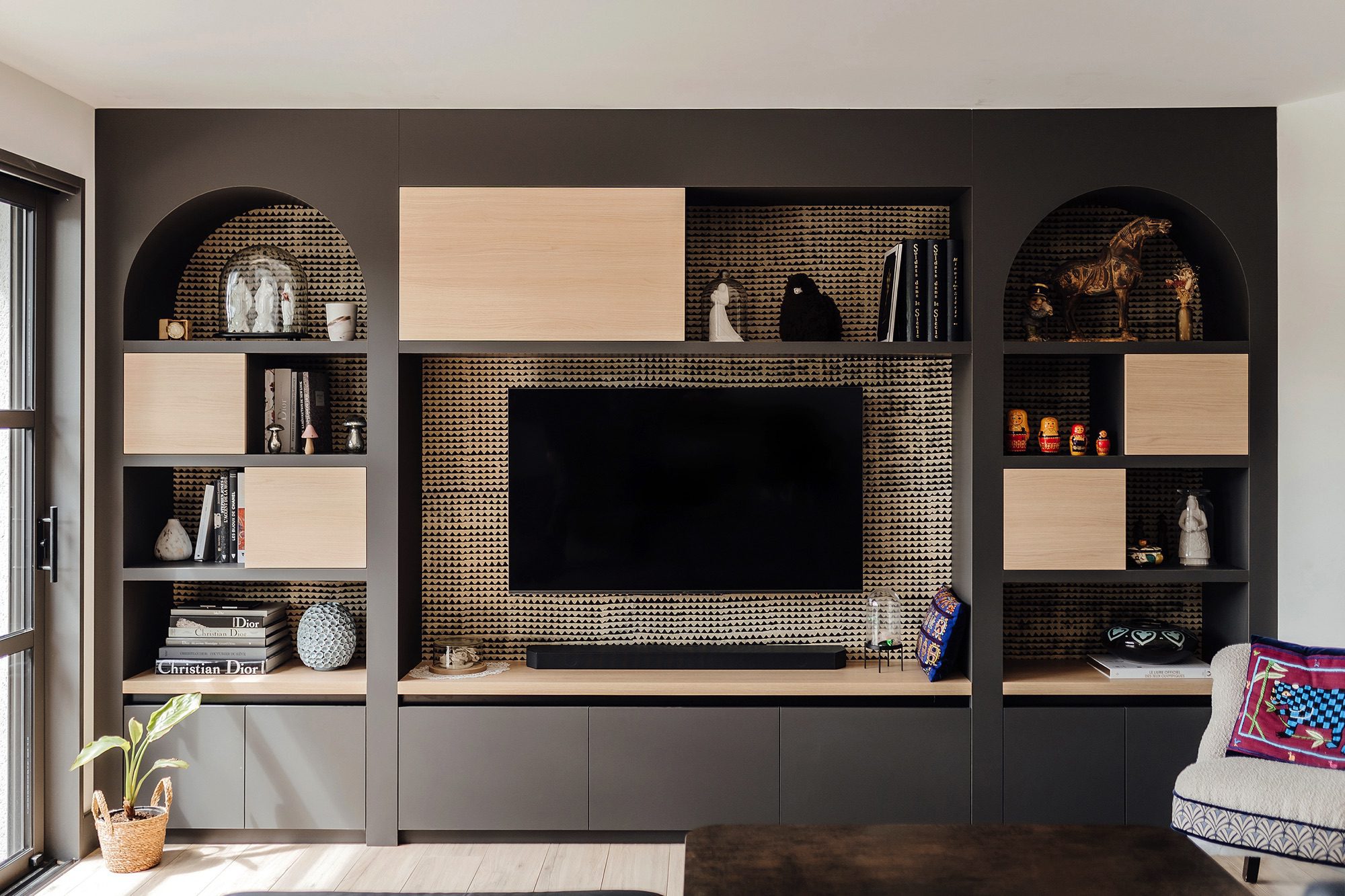 Bibliothèque sur-mesure en teintes de brun et beige avec un habillage papier peint à motif au fond. La niche centrale encadre la télévision. Sur les côtés, des colonnes en arche présentant des objets déco et des beaux livres.